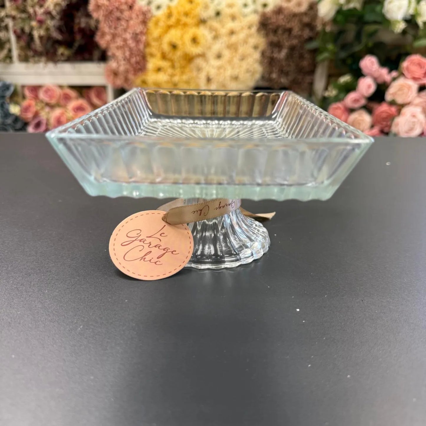 Clear glass candy dish on a black surface with a 'Garage Chic' tag in the foreground, flowers blurred in the background.