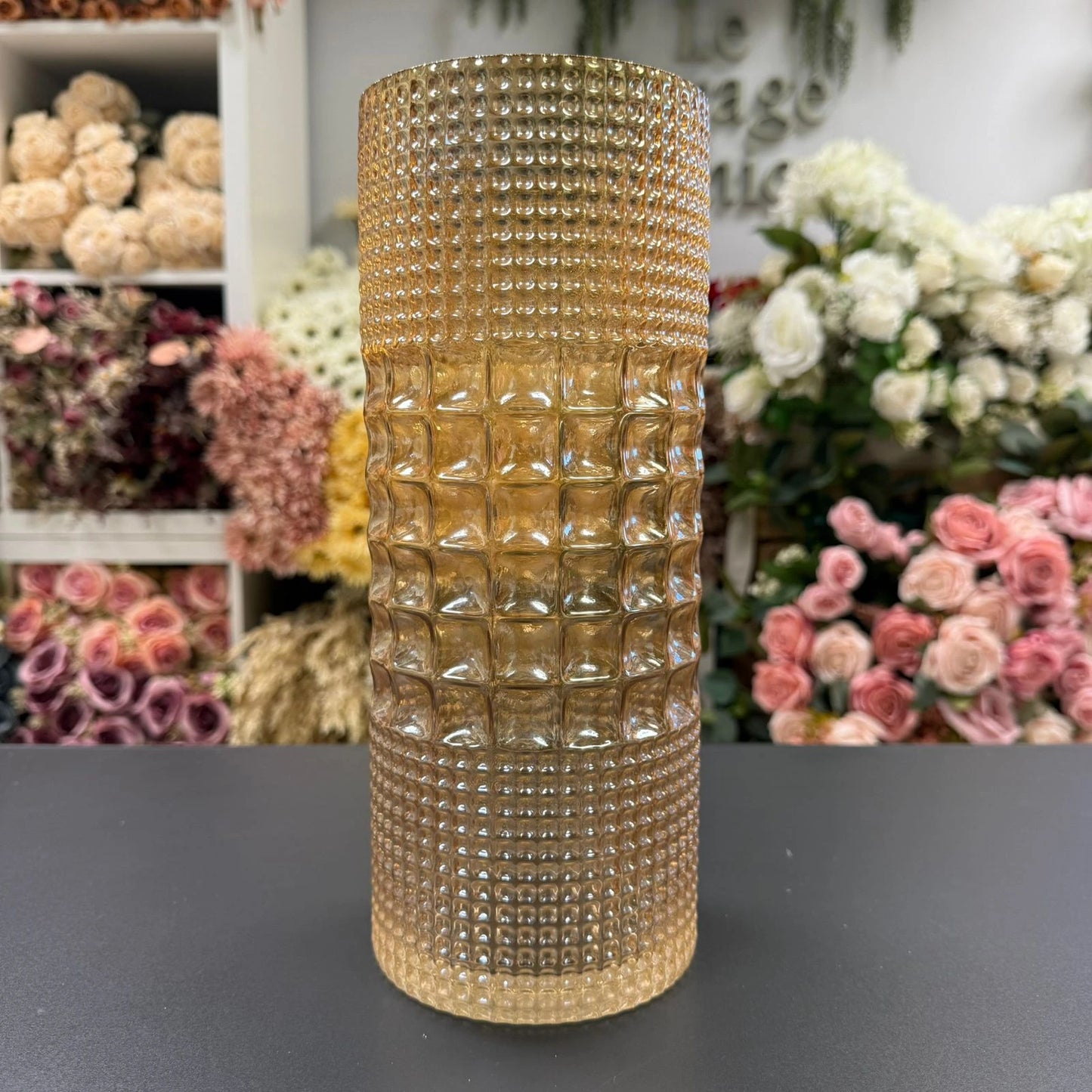 amber beaded vase on a dark surface with floral arrangements in the background