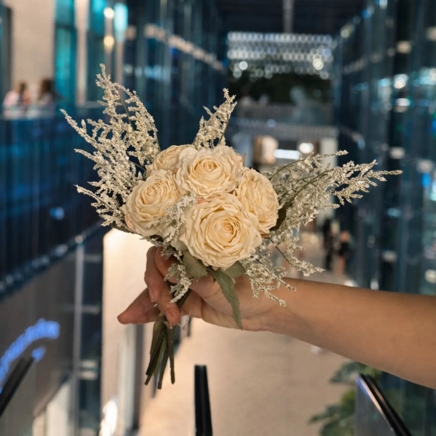 Hand holding a bouquet of beige roses with dried elements against a blurred indoor background