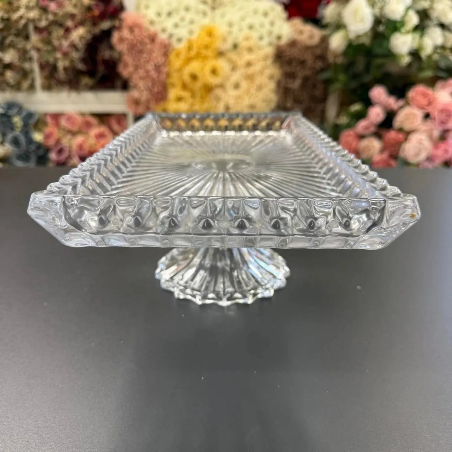 Decorative glass cake stand on a dark surface with flowers in the background