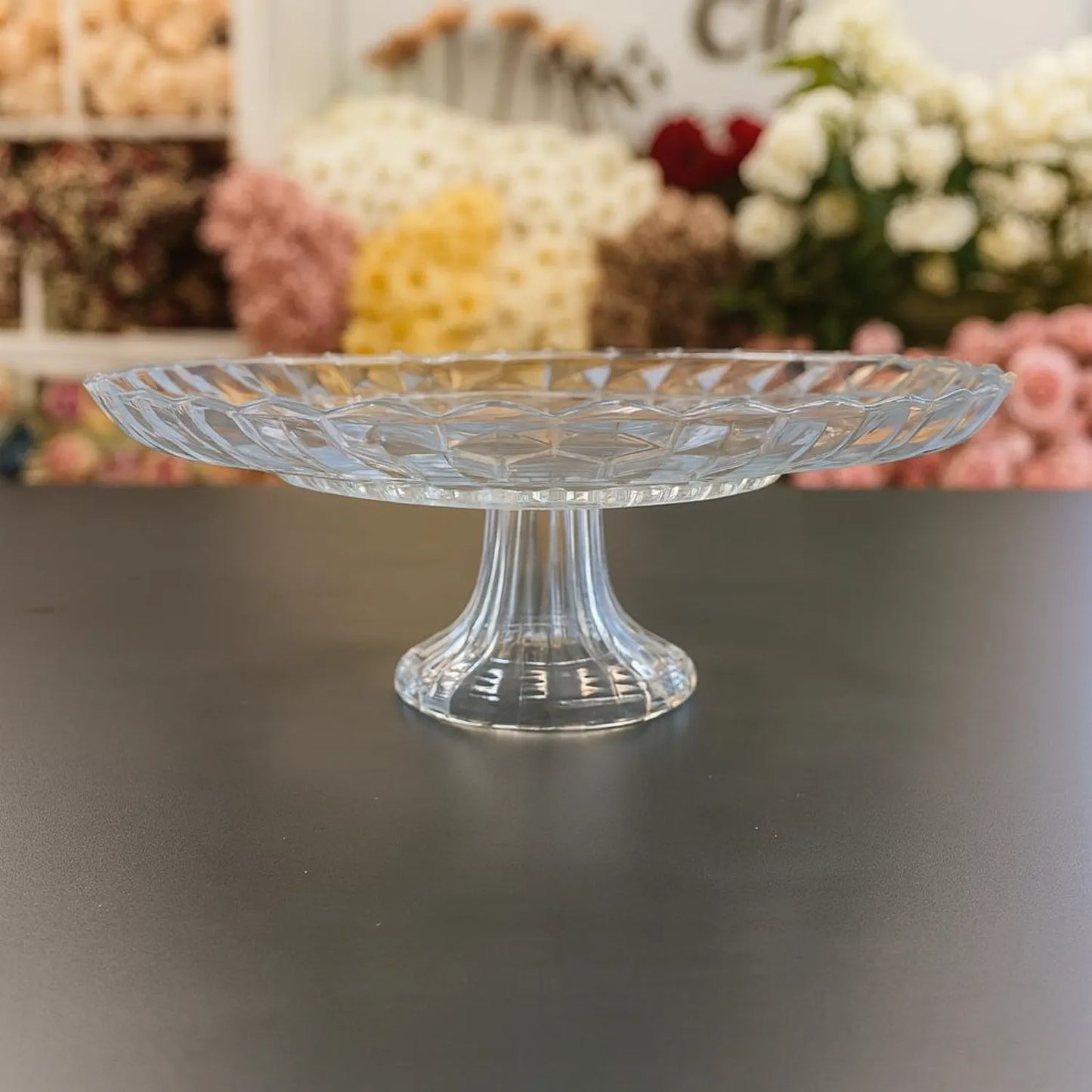 Clear glass cake stand on a dark surface with blurred floral decorations in the background