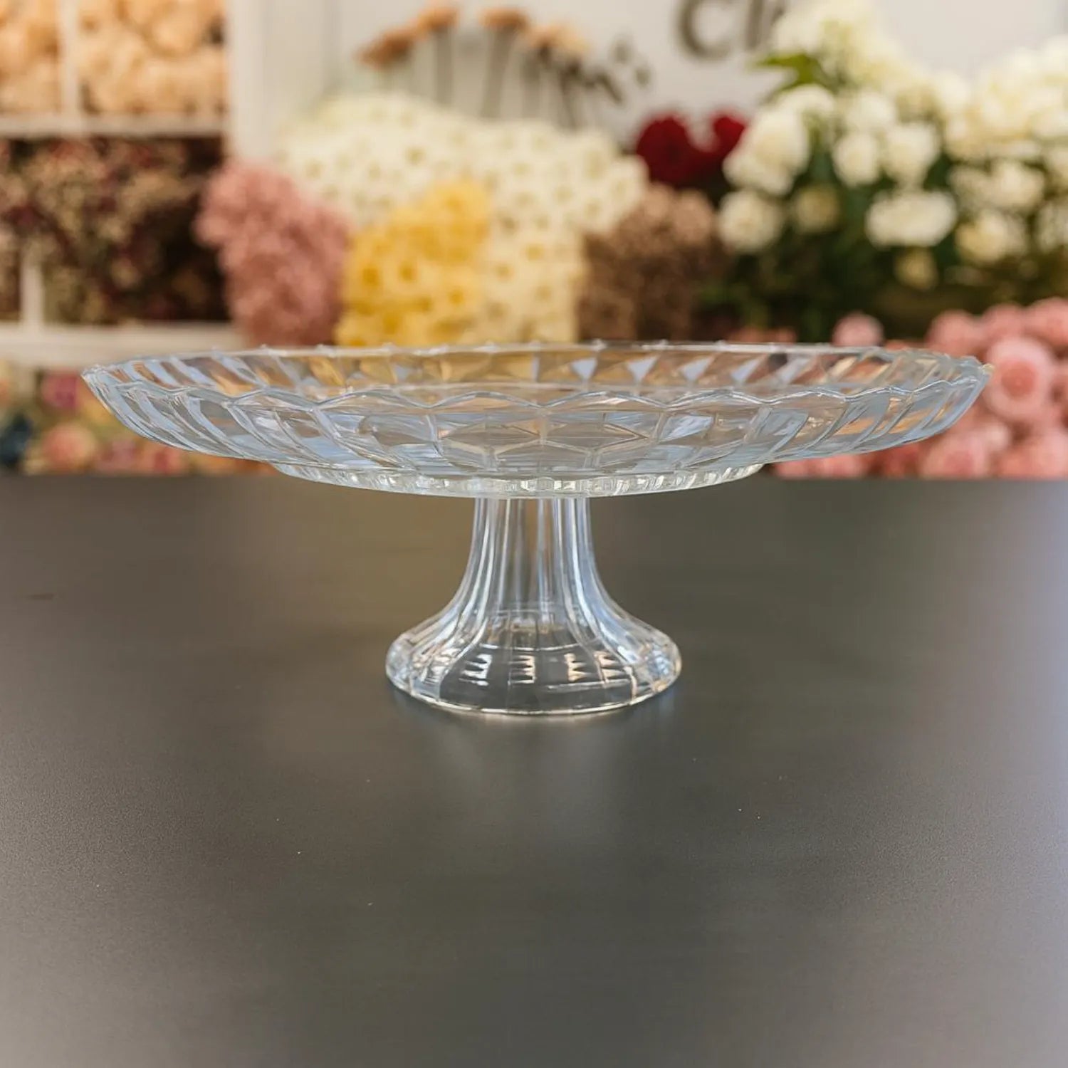 Clear glass cake stand on a dark surface with blurred floral decorations in the background