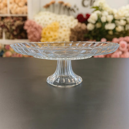 Clear glass cake stand on a dark surface with blurred floral decorations in the background