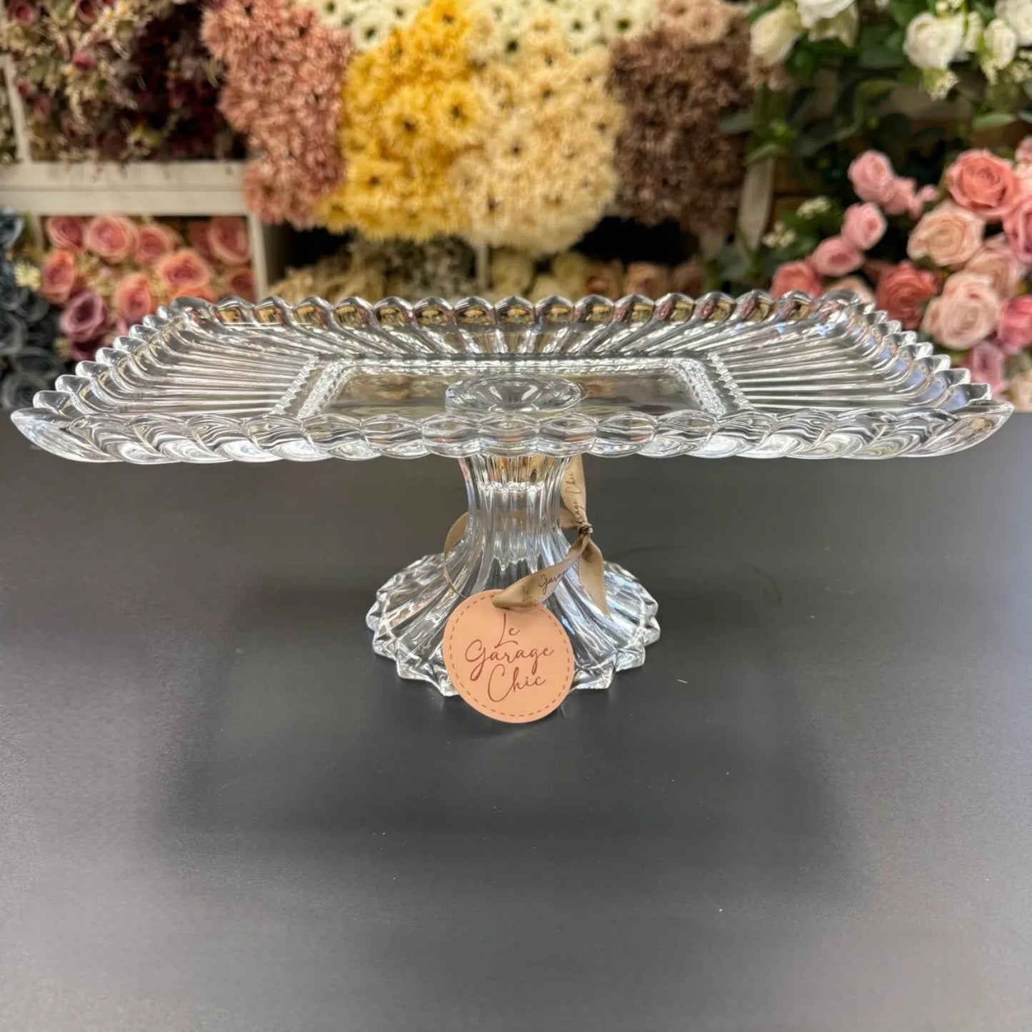 Clear glass cake stand with a decorative tag on a dark surface, flowers in the background