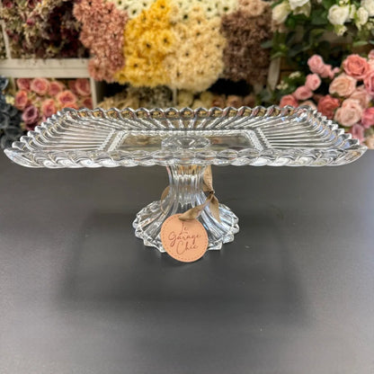 Clear glass cake stand with a decorative tag on a dark surface, flowers in the background