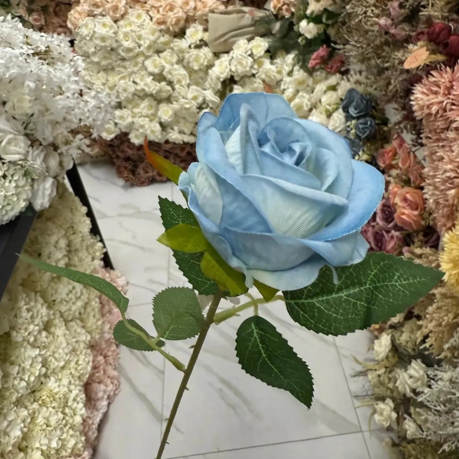 Blue artificial rose with green leaves against a background of various flowers.