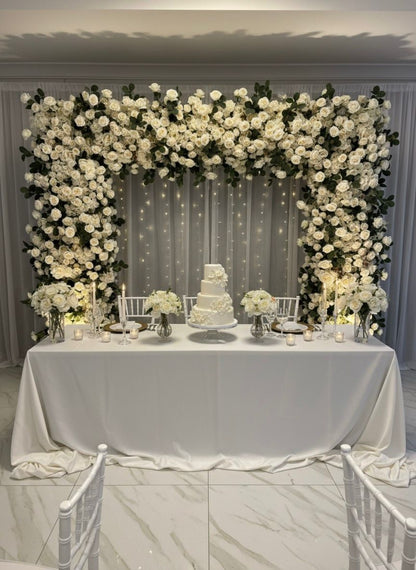 Decorative floral arch with a wedding cake on a table in a room.