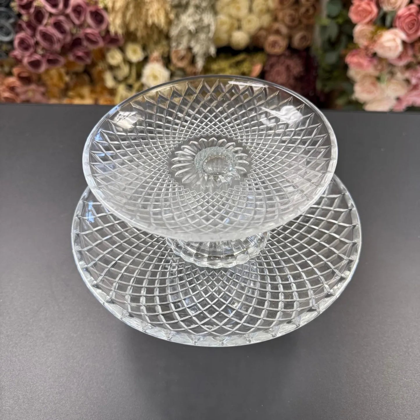 Two clear glass plates with textured design on a dark surface, floral decorations in the background.