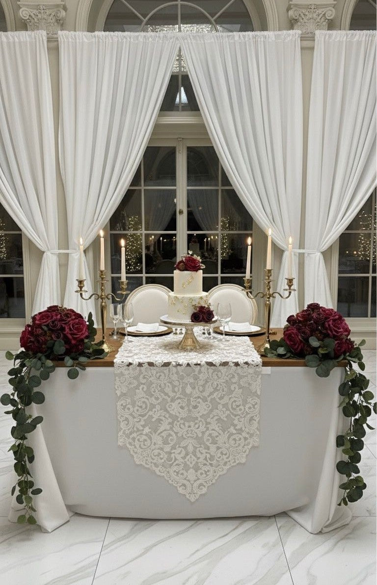 Decorated table with white lace runner, red flowers, and candles under large white curtains.