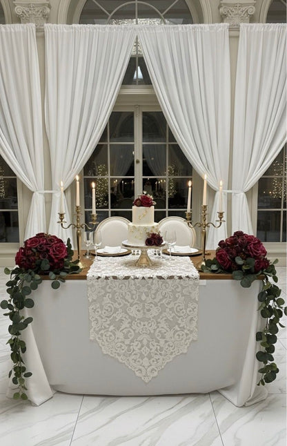 Decorated table with white lace runner, red flowers, and candles under large white curtains.