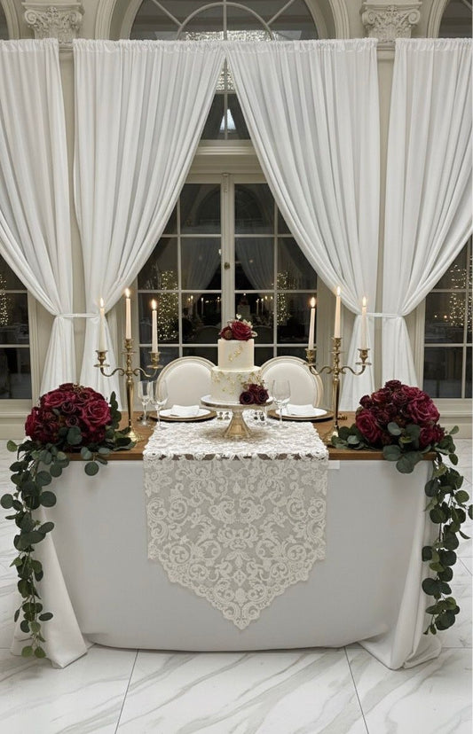 Decorated table with white lace runner, red flowers, and candles under large white curtains.