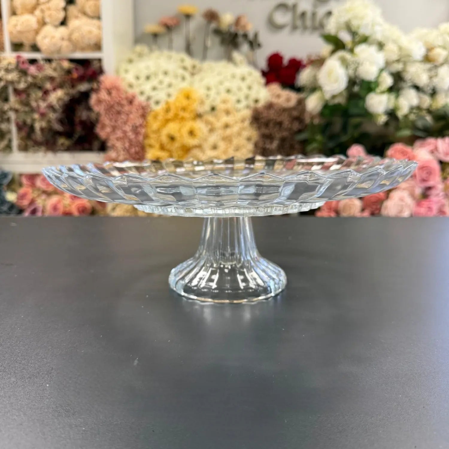 Clear glass cake stand on a dark surface with floral decorations in the background