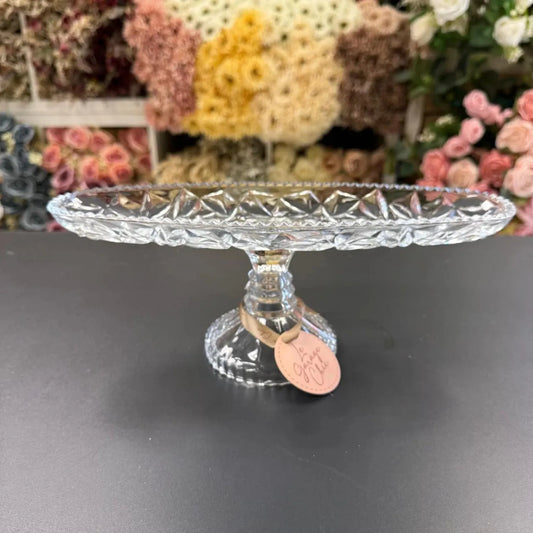 Clear glass cake stand with decorative base on a dark surface, flowers in the background