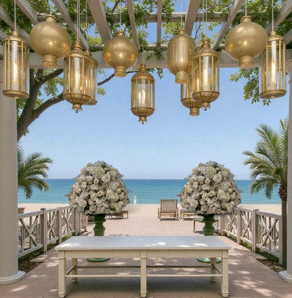 Outdoor setting with hanging gold lanterns, a table, and floral arrangements by the ocean.