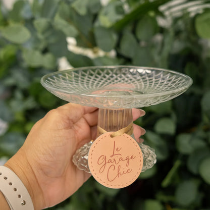 Hand holding a decorative glass plate with a 'Le Garage Chic' tag against a green leafy background