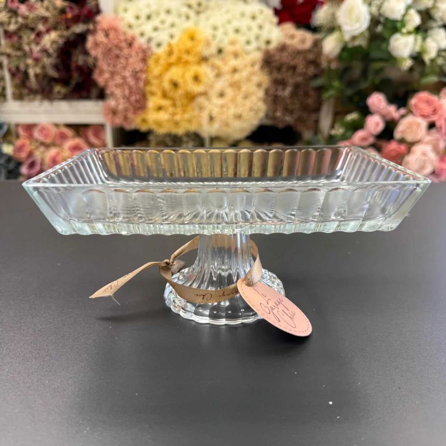 Clear glass footed dish with a tag on a dark surface, flowers in the background