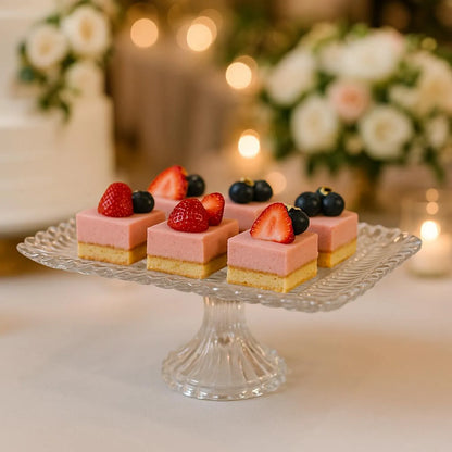 Small pink desserts with berries on a glass stand, blurred floral and candle background