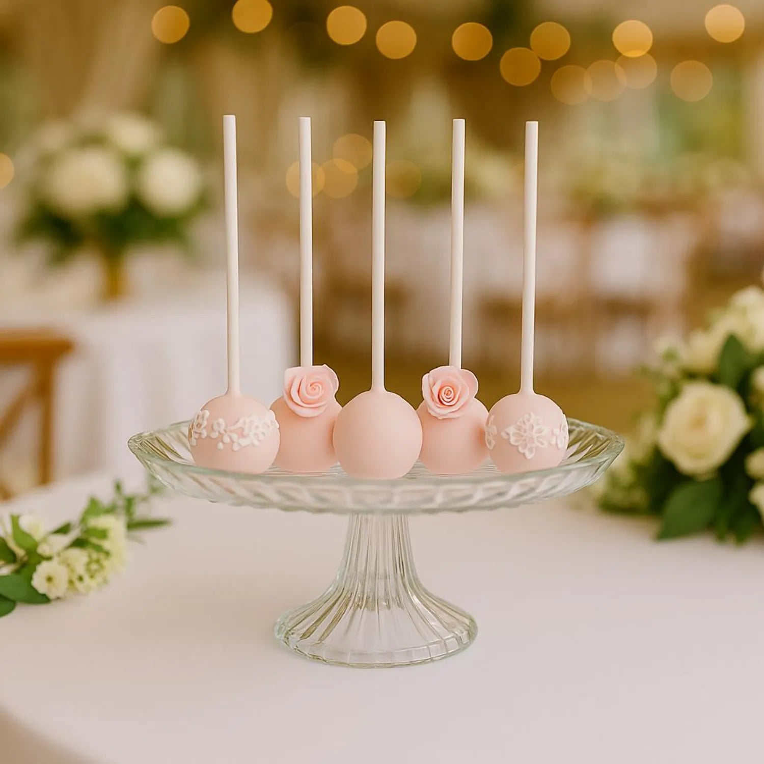 Decorative cake pops on sticks with flowers on a glass stand against a blurred background of flowers and lights.