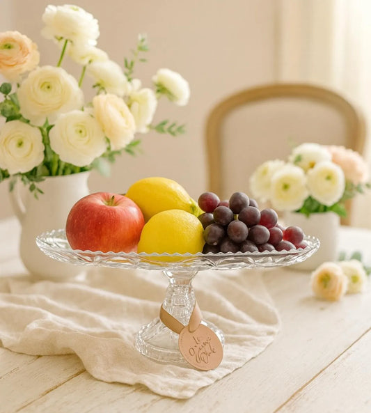 Fruit platter with apples, lemons, and grapes on a glass stand with flowers in the background.