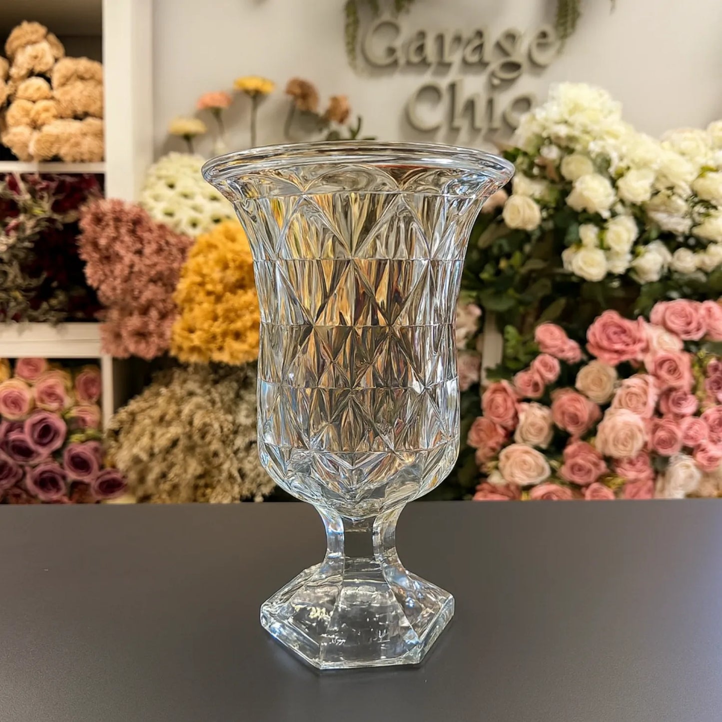Clear glass vase on a dark surface with floral arrangements in the background