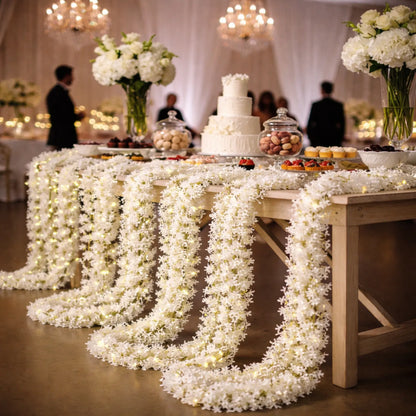 Decorated wedding cake table with floral arrangements and lights in a banquet hall.