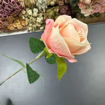 Single artificial rose with green stem on a gray surface, with a blurred background of flowers.