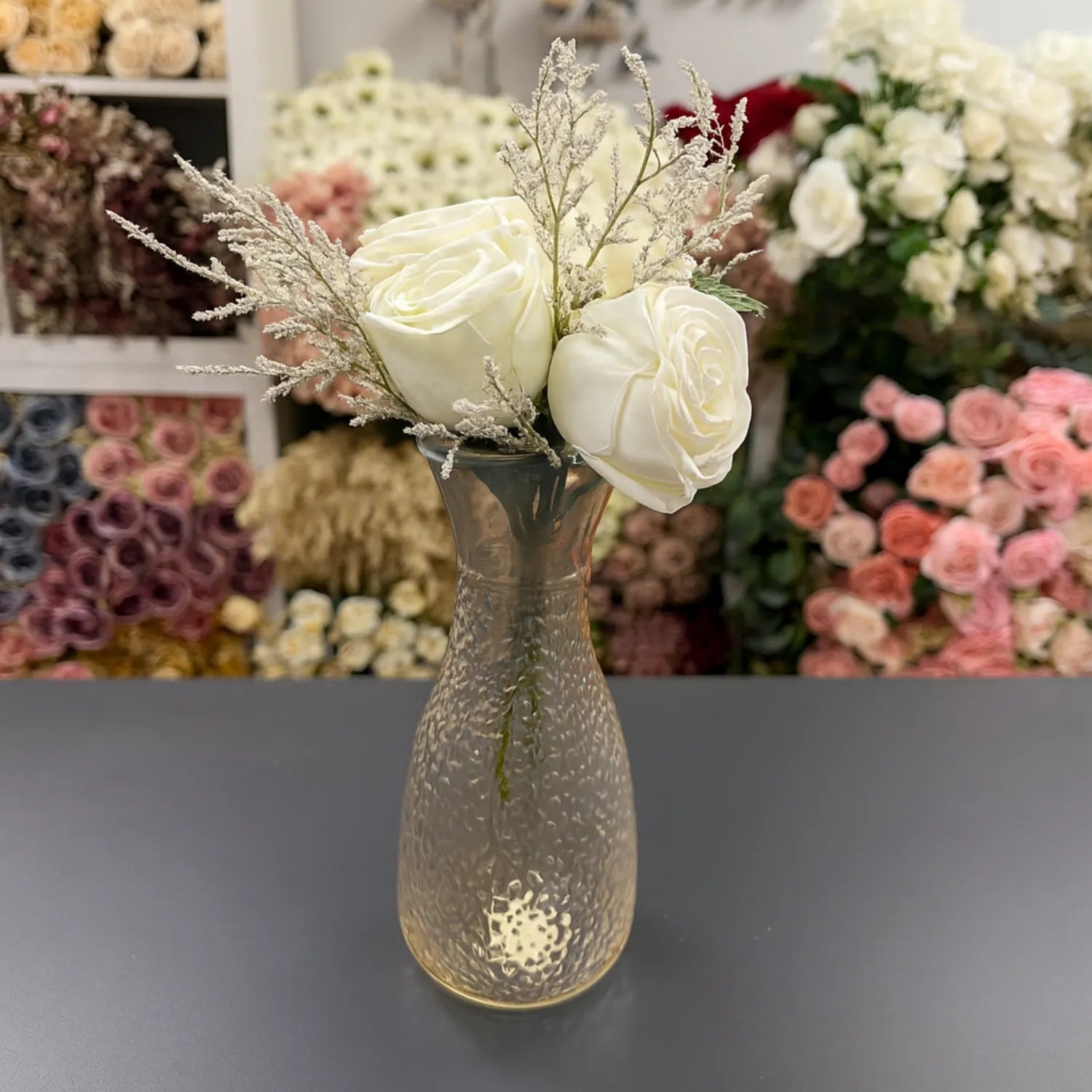Decorative vase with white flowers on a dark surface, with floral arrangements in the background.