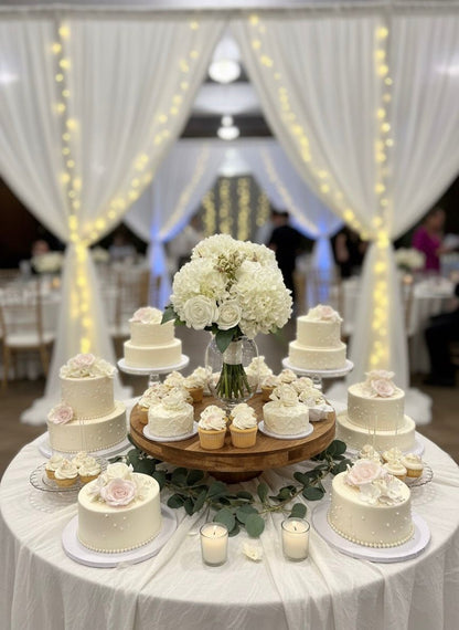 Decorative table with small white cakes and a centerpiece of white flowers, set against a draped backdrop with string lights.