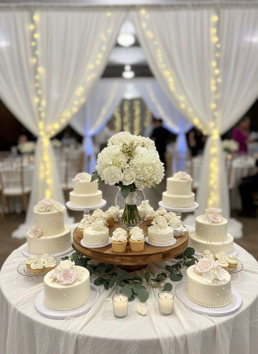 Decorative table with small white cakes and a centerpiece of white flowers, set against a draped backdrop with string lights.