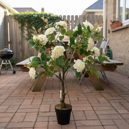 Potted artificial white flower plant on a patio with a wooden table and outdoor furniture in the background.